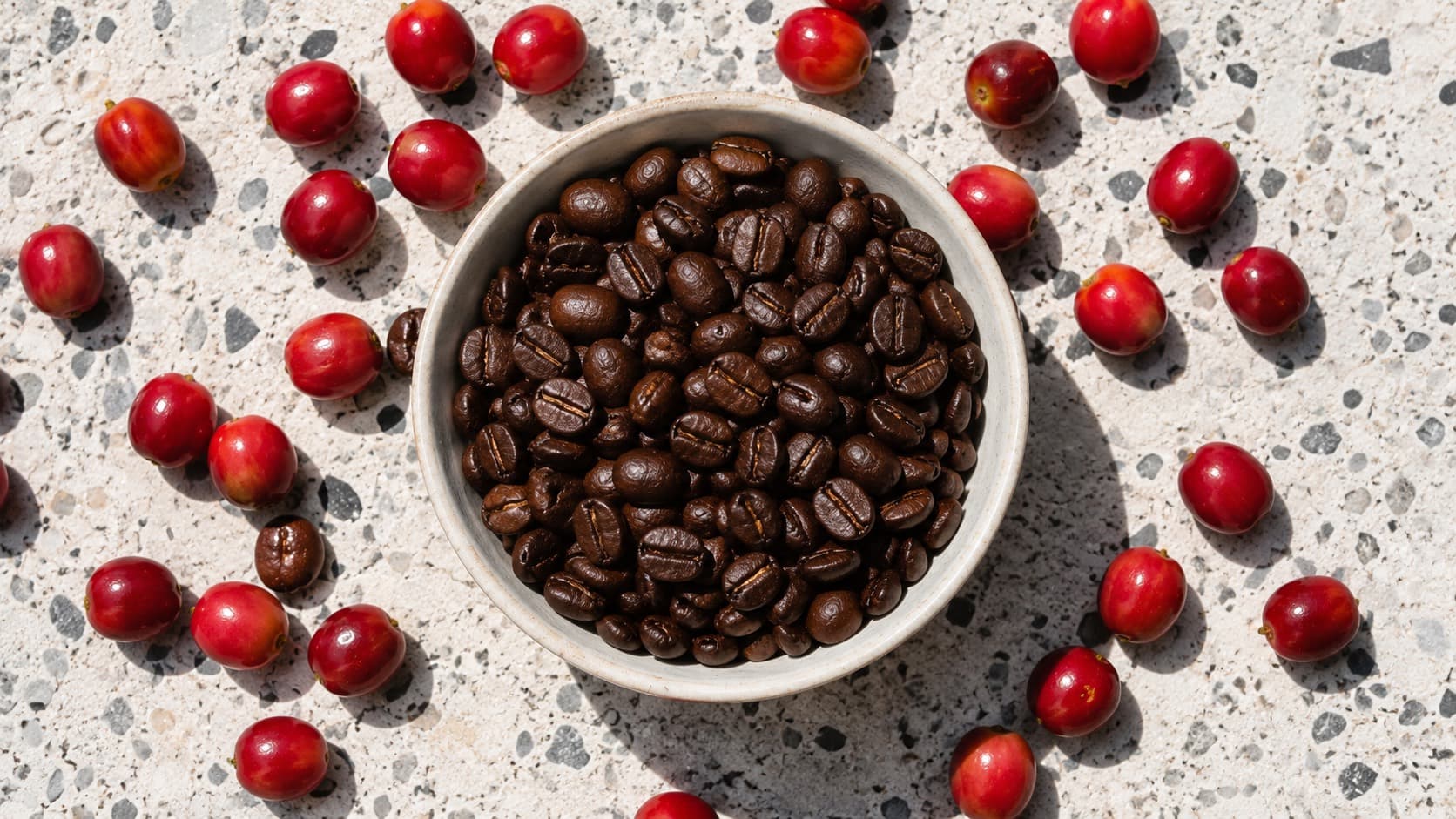 Dunkel geröstete Kaffeebohnen in einer Schale, umgeben von reifen roten Kaffeekirschen