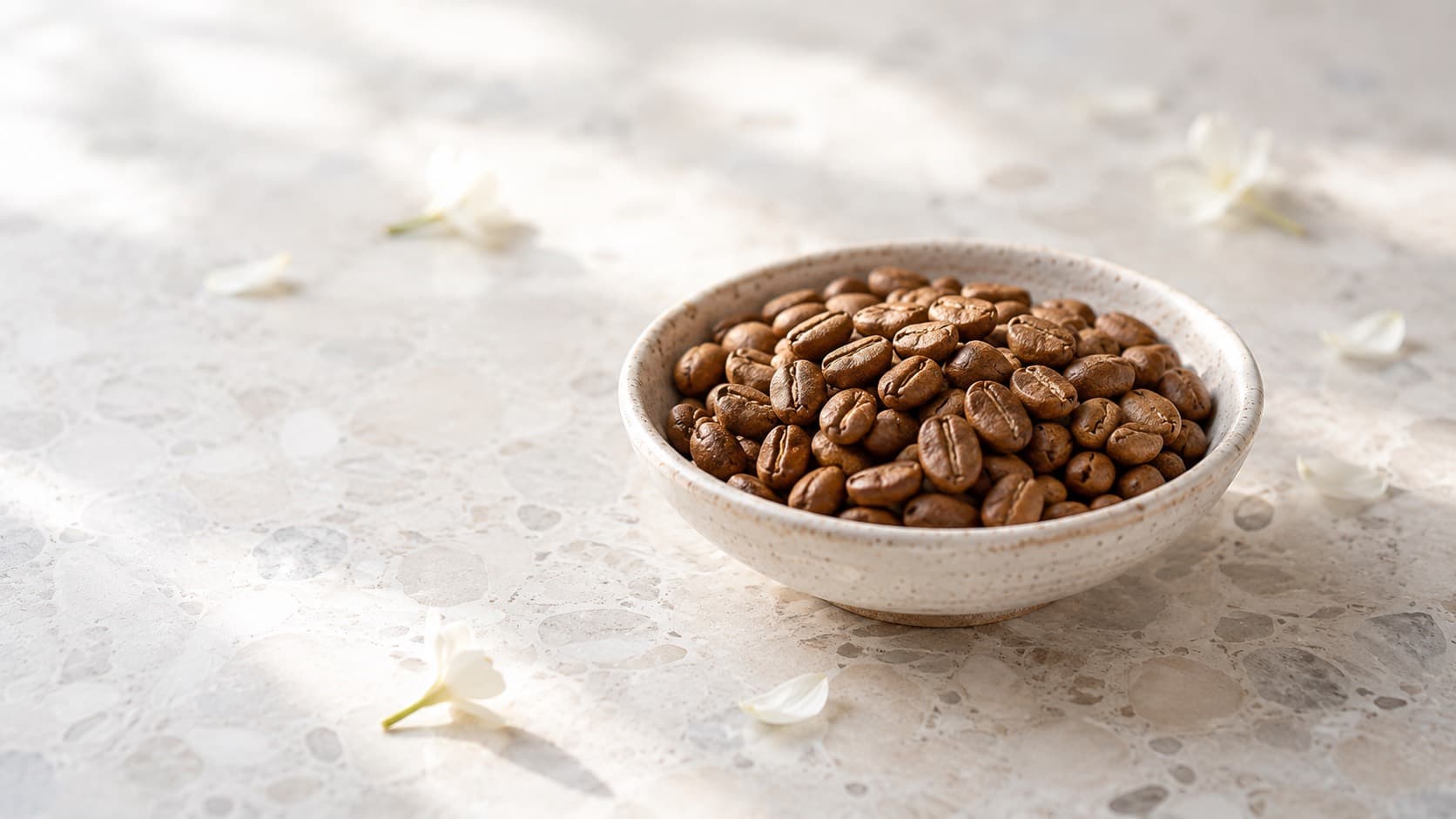 Mittelbraun geröstete Kaffeebohnen in einer hellen Keramikschale, daneben weiße Blüten auf einer Steinfläche